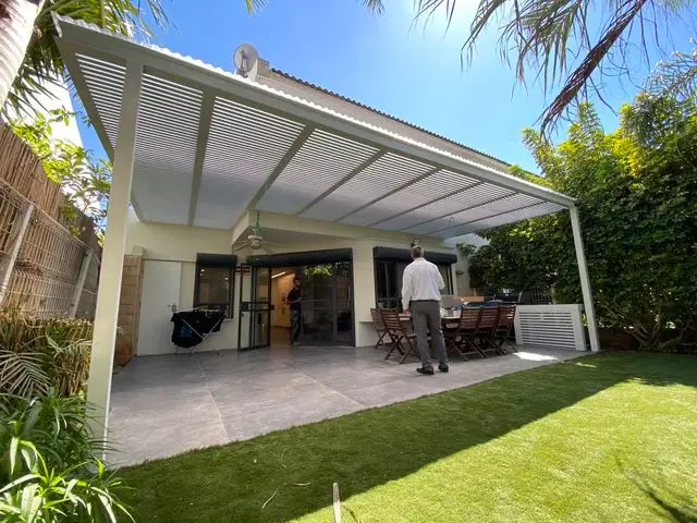 פרגולת אלומיניום לבנה על עמודים מעל פינת אוכל, מבט מהמדשאה אל הבית