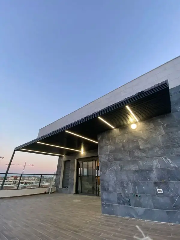פרגולת אלומיניום אנתרציט עם פסי תאורה מעל מרפסת גג בשעת שקיעה