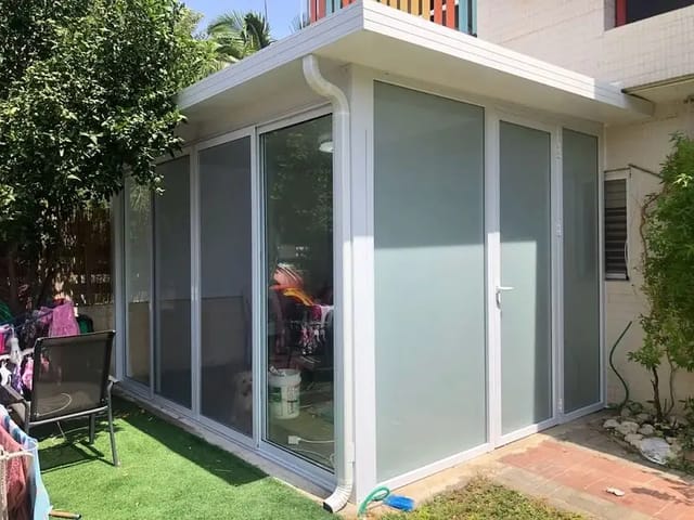 פרגולה סגורה לבנה צמודה לבית עם זכוכית חלבית, דלת ומרזב