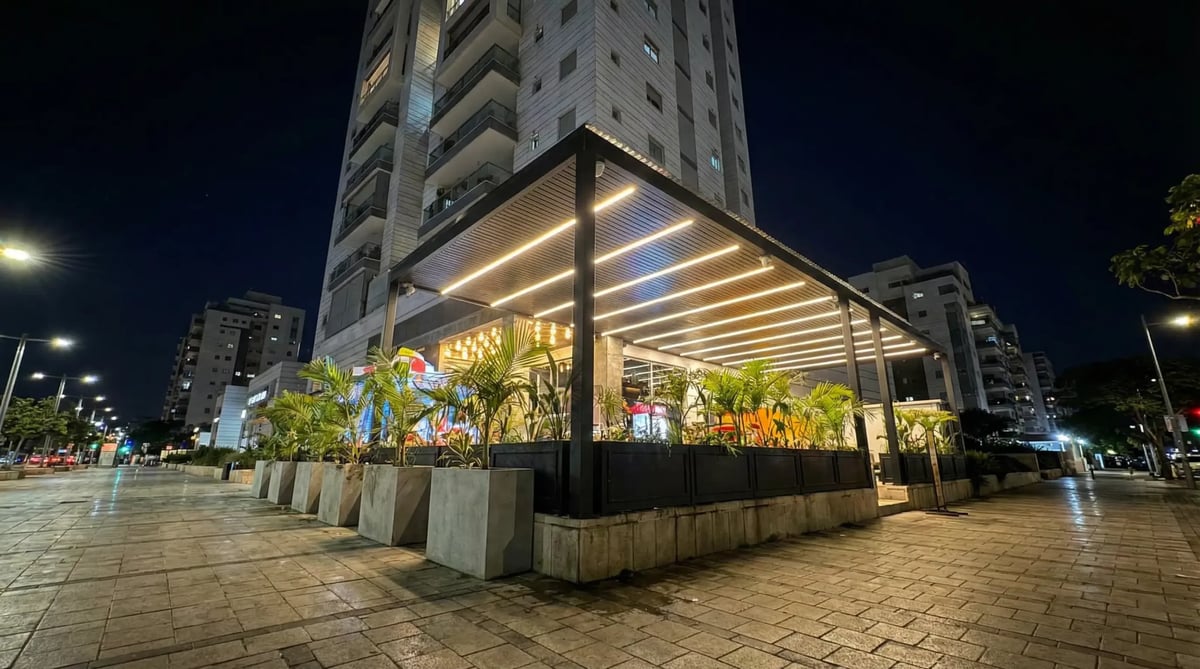 פרגולה פינתית שחורה עם תאורה ואדניות ברחבה מרוצפת לרגלי מגדל מגורים