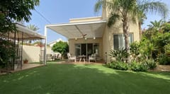 פרגולה לבנה צמודה לבית בקצה מדשאה, מבט מרוחק מהגינה