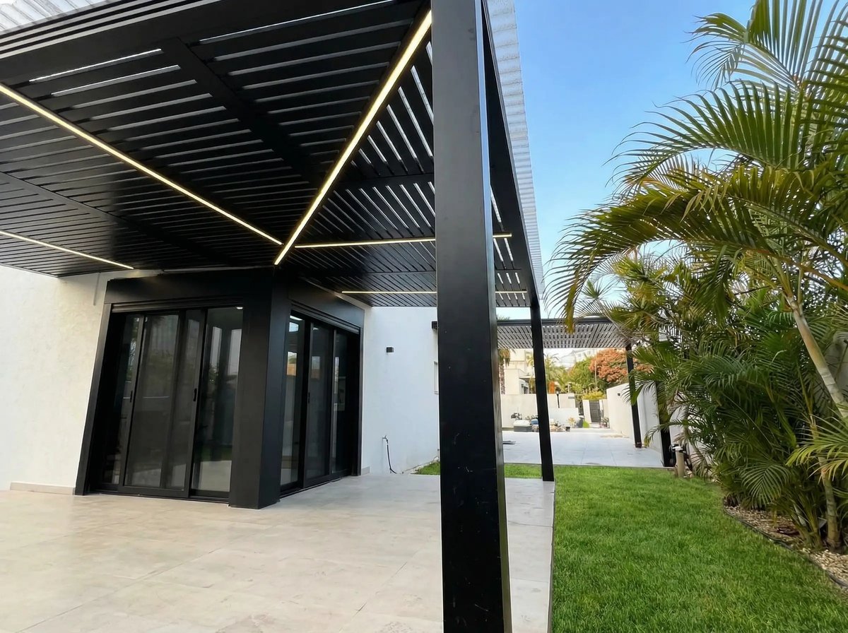 עמוד שחור ופסי תאורה בפרגולה מעל ריצוף, ליד מדשאה ודקלים