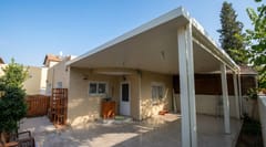 פרגולת קירוי מלא לבנה לאורך חזית הבית מעל חצר מרוצפת