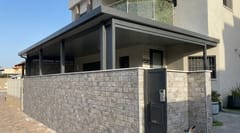 פרגולה אפורה עם קירוי אטום מעל חצר קדמית, מוקפת גדר מחופה אבן